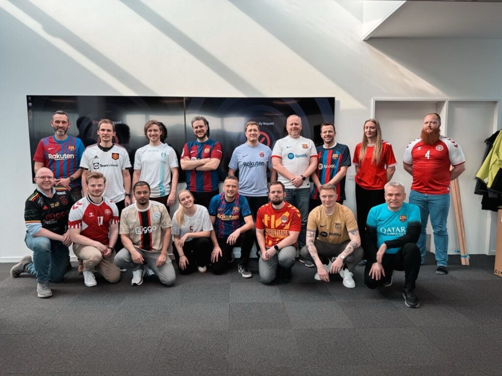 Group photo of adults in soccer jerseys posing indoors in front of a wall-mounted screen; diverse team smiling at camera. » admin by request