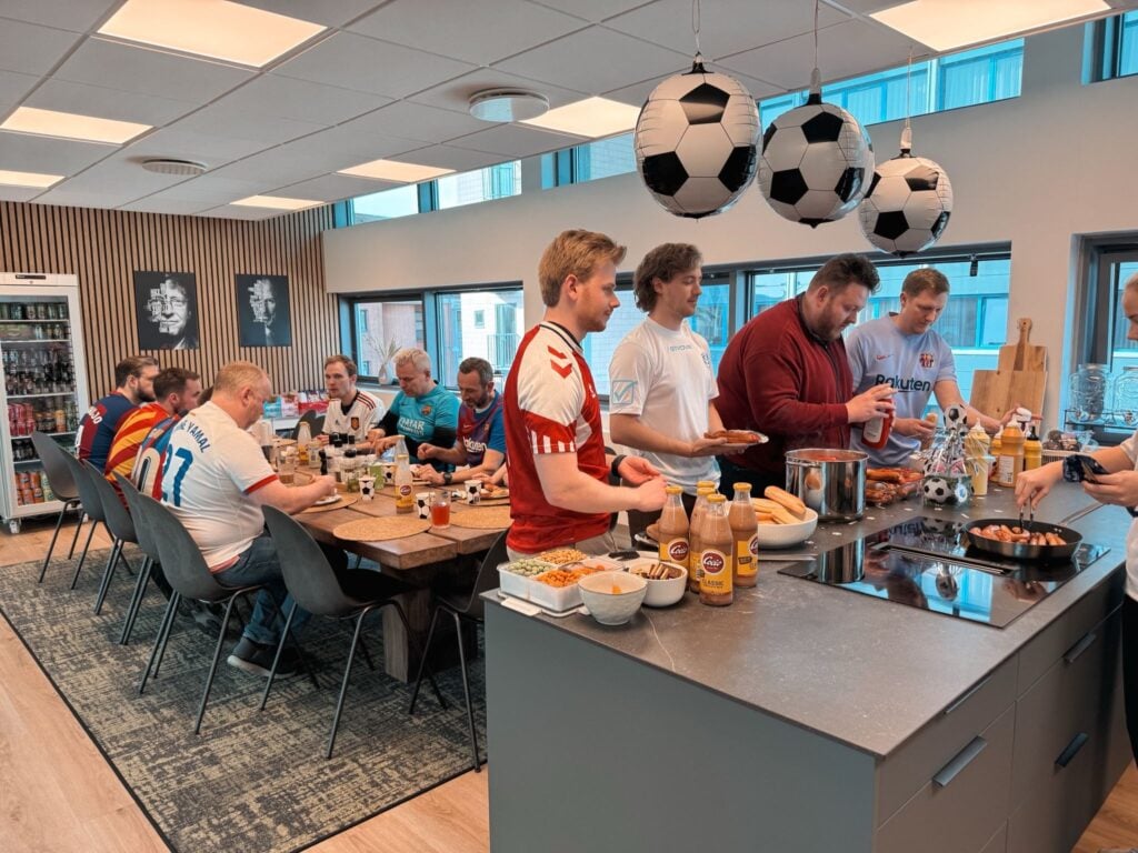 Group of men in soccer jerseys seated at a long table, enjoying a casual meal in a modern office break room with football-themed balloons overhead, including large hanging soccer balls over the counter. » admin by request