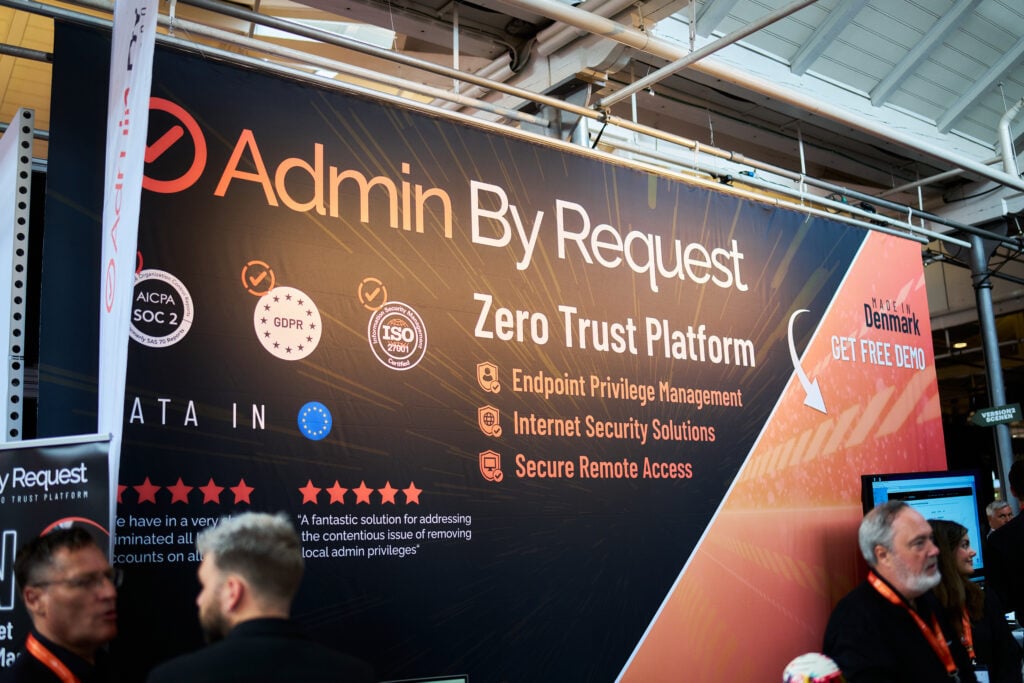 Wide banner reading 'admin by request' with 'zero trust platform' at a tech conference, supported by logos; people stand in front of the display. » admin by request