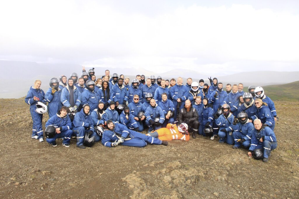 Group of people in blue coveralls and helmets posing on a rocky hilltop, one person in an orange suit lying in front. » admin by request