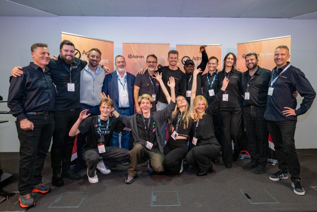 A large group of people posing together for a photo at an event, smiling and with arms around each other in front of orange banners with logos. » admin by request