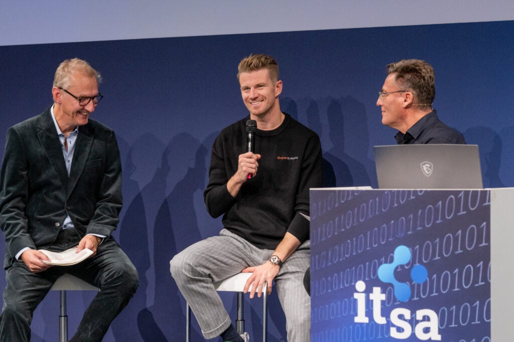 Three men seated on a stage in a panel discussion, the center man holding a microphone and smiling as the others listen. Blue backdrop with a tech-themed display on the right. » admin by request
