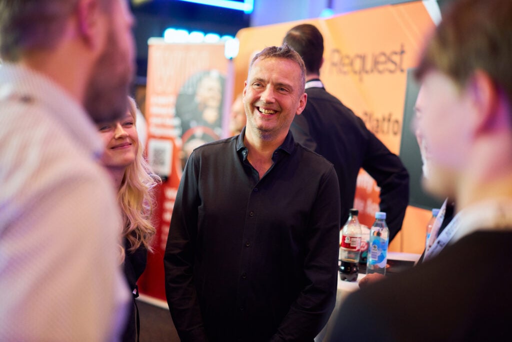 Smiling man in a black shirt stands at a busy booth, talking with people at an event. » admin by request » admin by request
