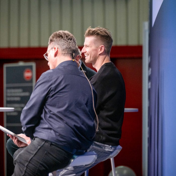 Two men seated on stools at a panel, smiling and talking, one wearing glasses and a headset mic in a red-walled venue. » admin by request