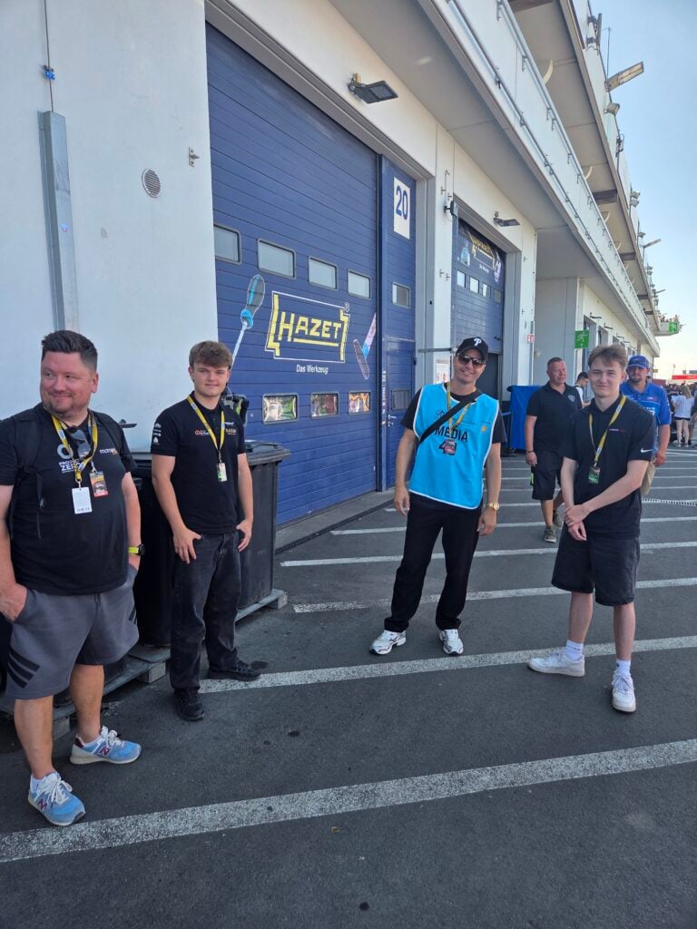 Group picture » admin by request Group of five men standing and posing in a parking lot in front of a large blue industrial garage door with a hazet logo and a '20' sign nearby, some wearing lanyards and badges. » admin by request