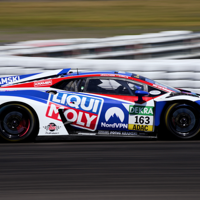 Blue, white, and red race car speeding on a track with sponsor decals visible (163 adac) and a blurred barrier in the background. » admin by request