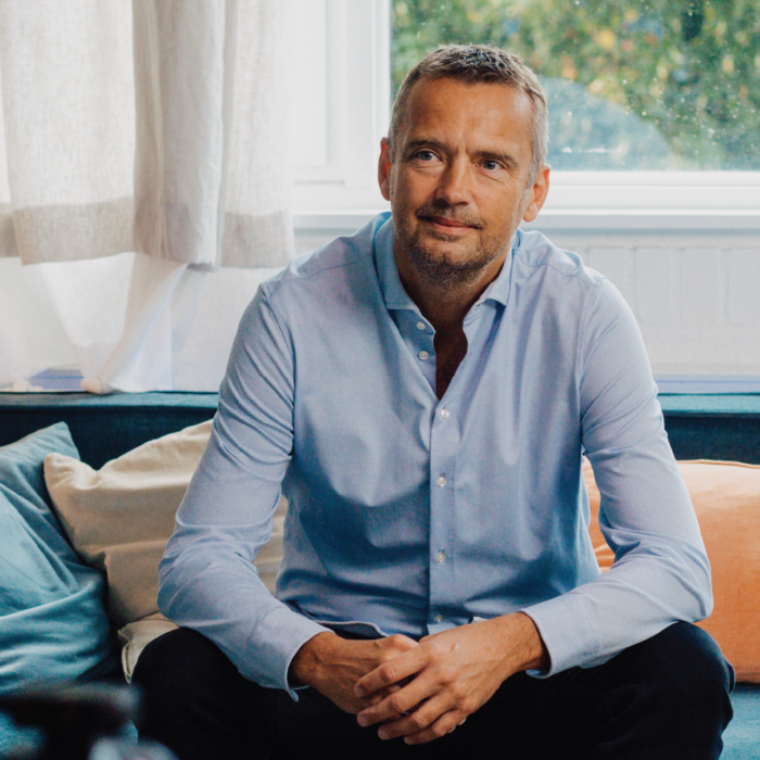 Man in a light blue button-down shirt sits on a blue sofa with teal and orange cushions in a bright living room by a window. » admin by request
