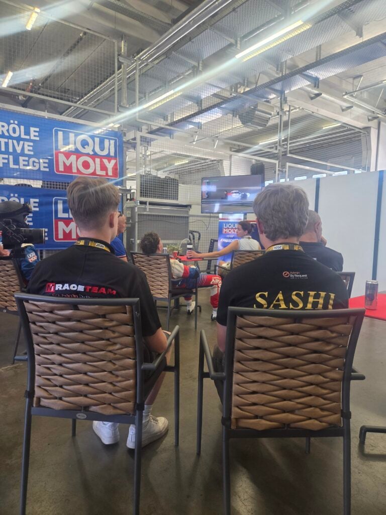 Luca and marcus » admin by request Audience seated in a workshop-like space watching a car video on a wall monitor, with liqui moly banners visible nearby. » admin by request