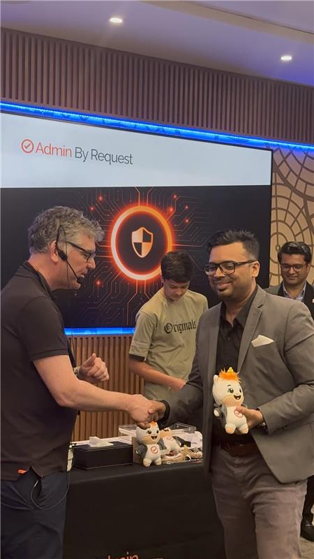 Image » admin by request Two men shake hands at a tech conference booth for admin by request; the man in a suit holds a plush unicorn while others look on. » admin by request