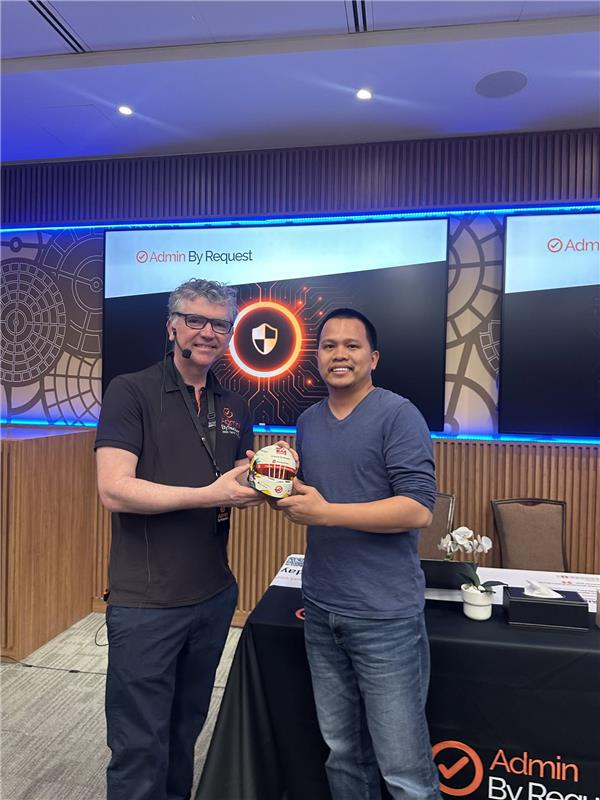 Image » admin by request Two men stand on a stage exchanging a small circular trophy, with branded screens in the background reading 'admin by request'. » admin by request