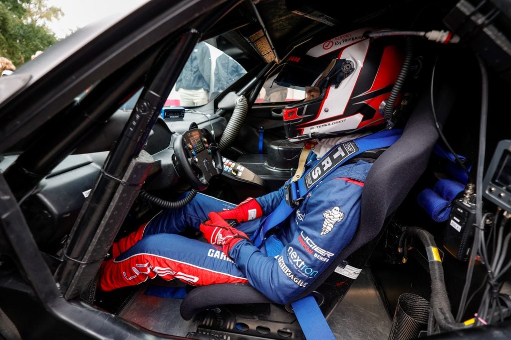 Ligier european series - spa heat - 21-23 august 2026 » admin by request Racing driver in cockpit wearing red-white helmet and blue suit, gripping steering wheel with visible control panel nearby. » admin by request