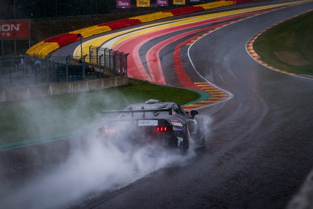 Ligier european series - spa heat - 21-23 august 2026 » admin by request Race car in a sharp bend on a wet circuit, spraying water as it powers through a rain-soaked corner with colorful red, yellow, and gray curbs nearby. » admin by request