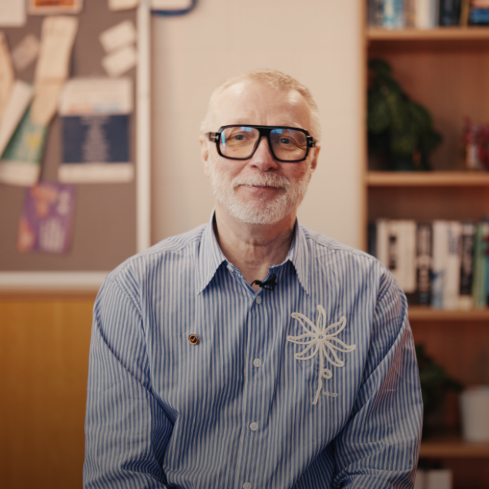 Older man with gray hair and beard, wearing glasses and a blue striped shirt, sits in a wood-paneled office with a bookshelf behind him. » admin by request