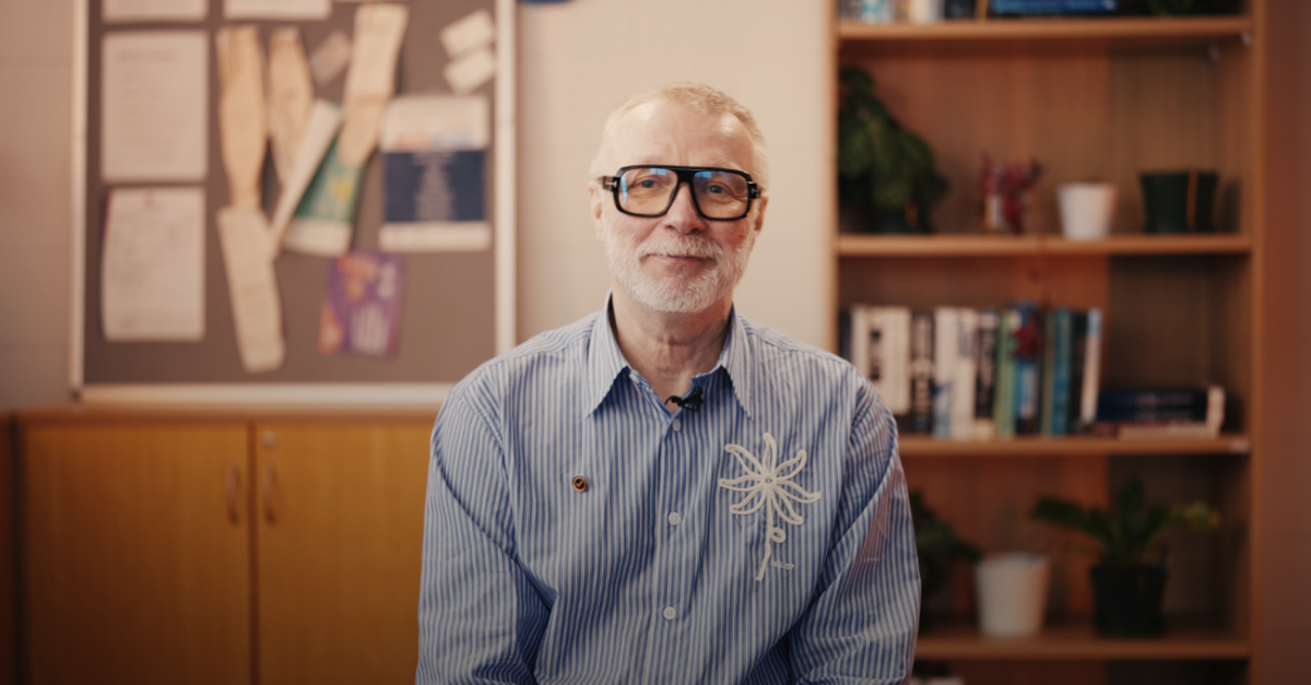 Older man with gray hair and beard, wearing glasses and a blue striped shirt, sits in a wood-paneled office with a bookshelf behind him.