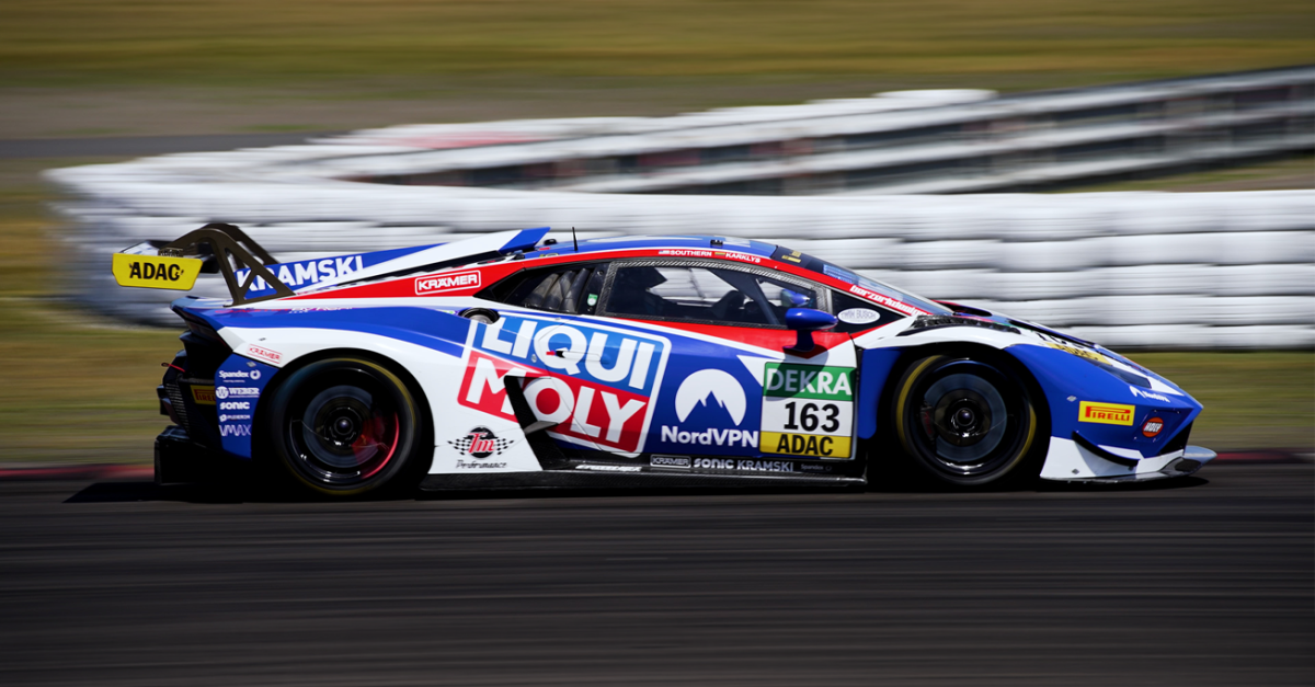 Blue, white, and red race car speeding on a track with sponsor decals visible (163 ADAC) and a blurred barrier in the background.