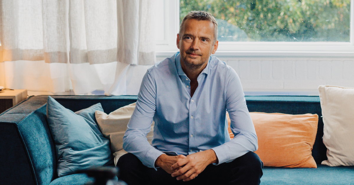 Man in a light blue button-down shirt sits on a blue sofa with teal and orange cushions in a bright living room by a window.