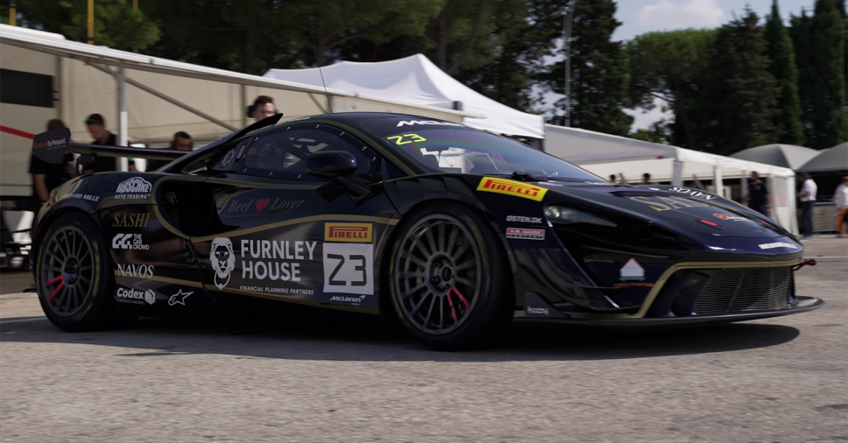 Black race car with number 23 and sponsor logos in a paddock area under white tents, rear wing visible behind the vehicle.