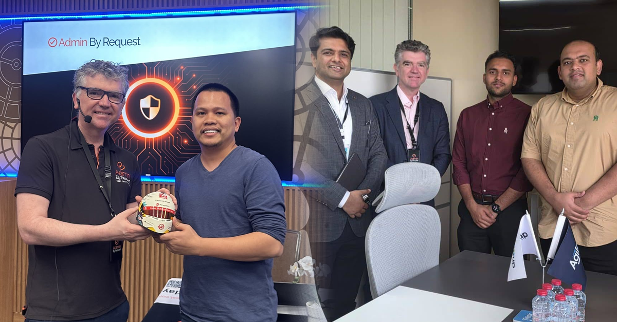 Two men exchange a small branded ball trophy in an office event, with a large 'Admin By Request' screen behind them and colleagues nearby.