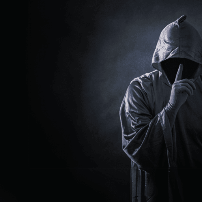 Hacker in a hospital gown and hood holding his finger up to his mouth as if to say, 'shh'. For an admin by request blog on ransomware attacks in the healthcare sector » admin by request