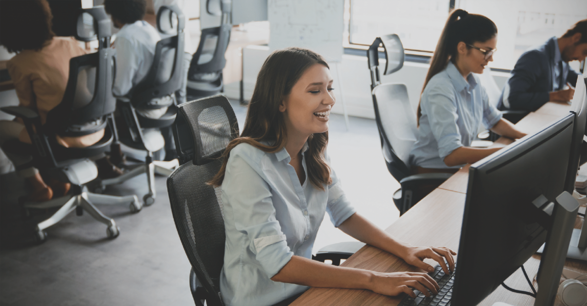 A happy woman on her computer with four of her co-workers in the background of the office.